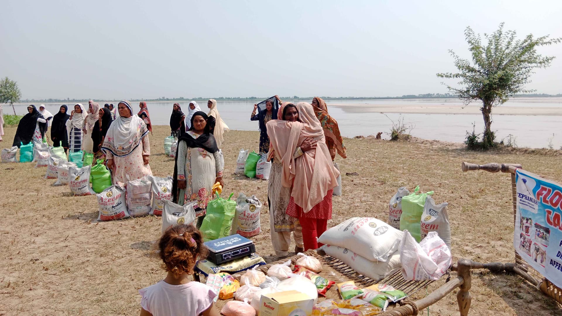 Sister Zeph, a woman dressed in red and light pink, hugs a woman while distributing grocery packs. Other women and a girl stand nearby, surveying the flooded horizon.