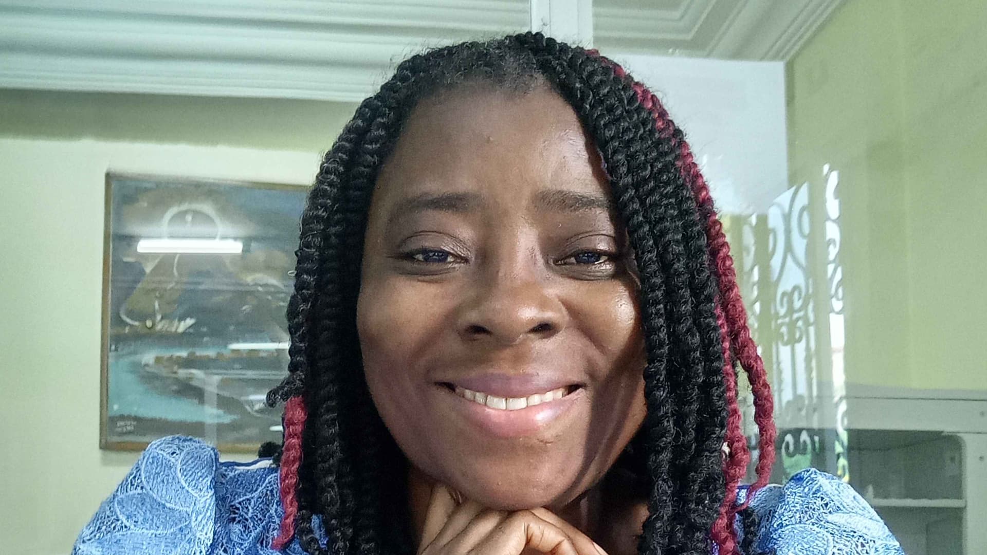 I am sitting on my office desk, with a smile, hand on my chin, wearing a blue white dress and hair braided in twist with black and a touch of red.