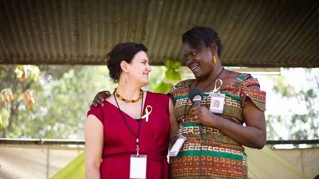 Photo of Jensine and Leah in colorful patterned clothes standing in Kenya under trees. Leah holds microphone and they are looking into each others eyes with deep friendship and connection
