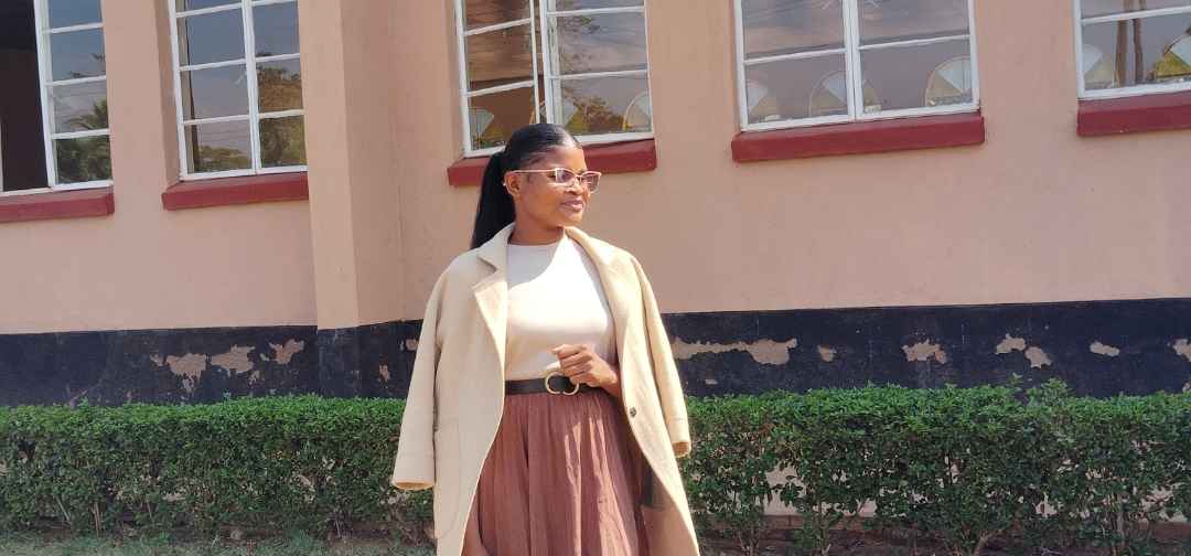 A young woman standing outdoors in front of a building, representing leadership, hope, and commitment to empowering girls through education, health, and community development.