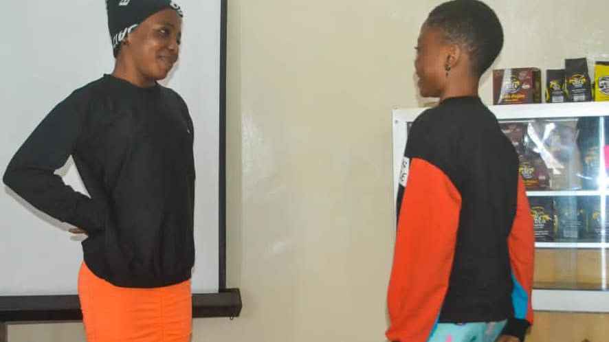 Two adolescent girls standing face to face with each other, one wearing a black shirt over an orange pants and the other wearing a black and orange shirt over a sky blue flourish pants.