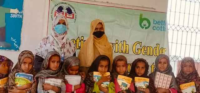 this image is showing the field facilitator and a teacher along with small girls who enrolled in school on day one and back side banner is showing logo of REEDS Pakistan left hand side and Better cotton logo right hand side