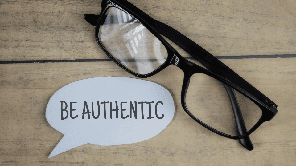 Image of a paper written "be authentic" next to it is a pair of glasses with a black frame. The background is a wooden beige.
