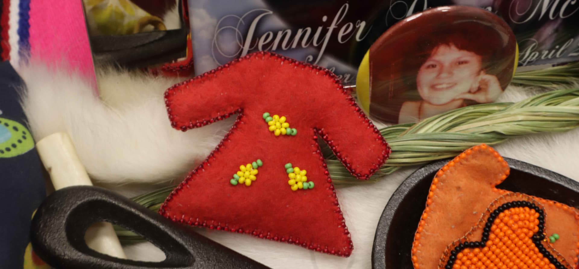 A miniature red dress pin with beads representing Gerri-Lee Pangman, the workshop facilitator's loved ones sits on a memorial altar alongside a portrait of Jennifer Dawn McPherson, a braided strand of sweetgrass, and a beaded orange shirt