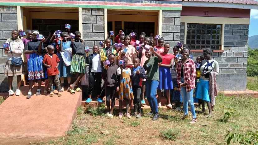 A group of girls in colorful outfits smile outside of a stone building during a menstrual awareness event. They hold boxes of menstrual hygiene supplies.