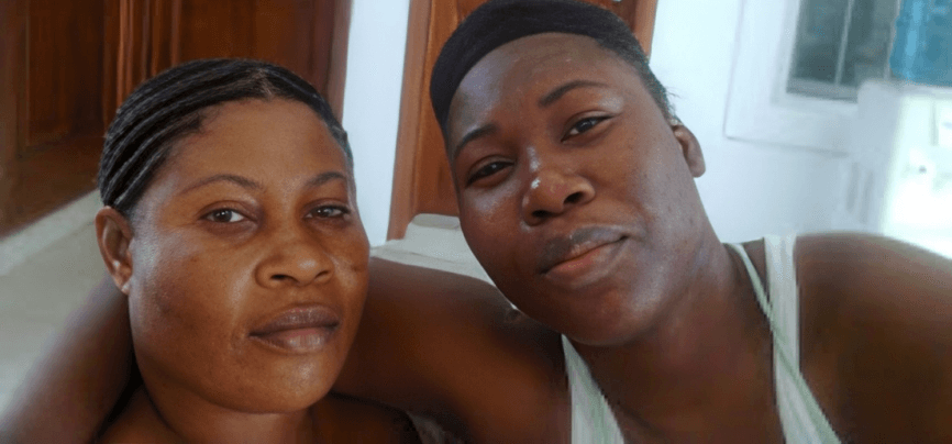 A mother and daughter sit closely together indoors, taking a selfie at home.