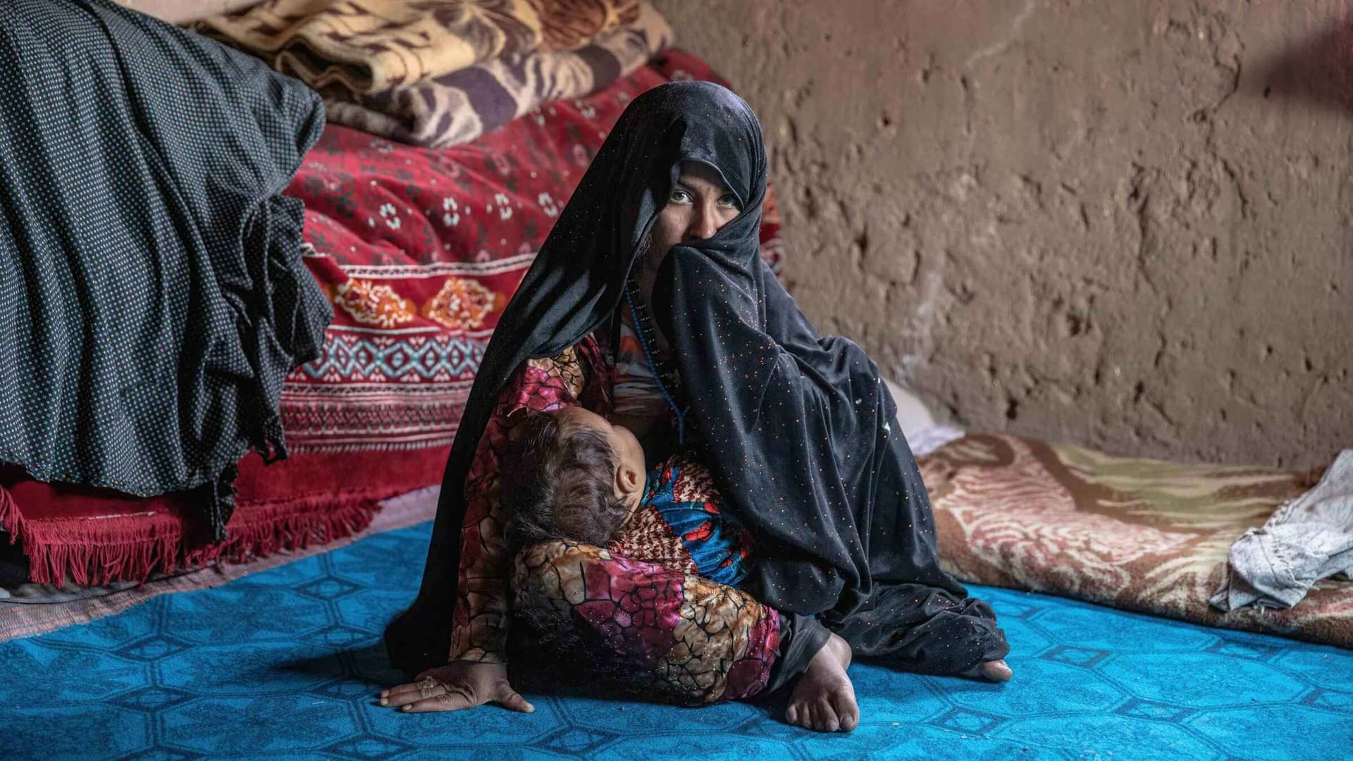 A woman in a hijab half-covers her face, sitting on a blue carpet inside her home.