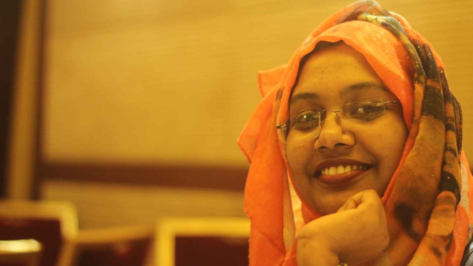 Mosfeka Zannat is photographed smiling with her hand folded under her chin. She has brown skin and glasses, wearing an orange patterned headscarf and silver watch on her wrist. Behind her are brown chairs and a wall.