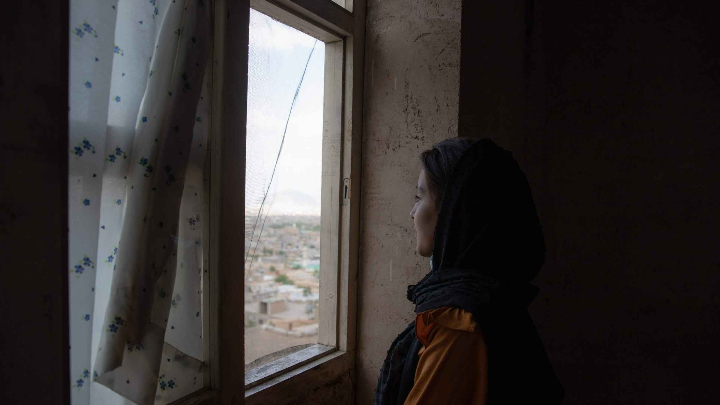 A woman looks out her window from the darkness of her home.