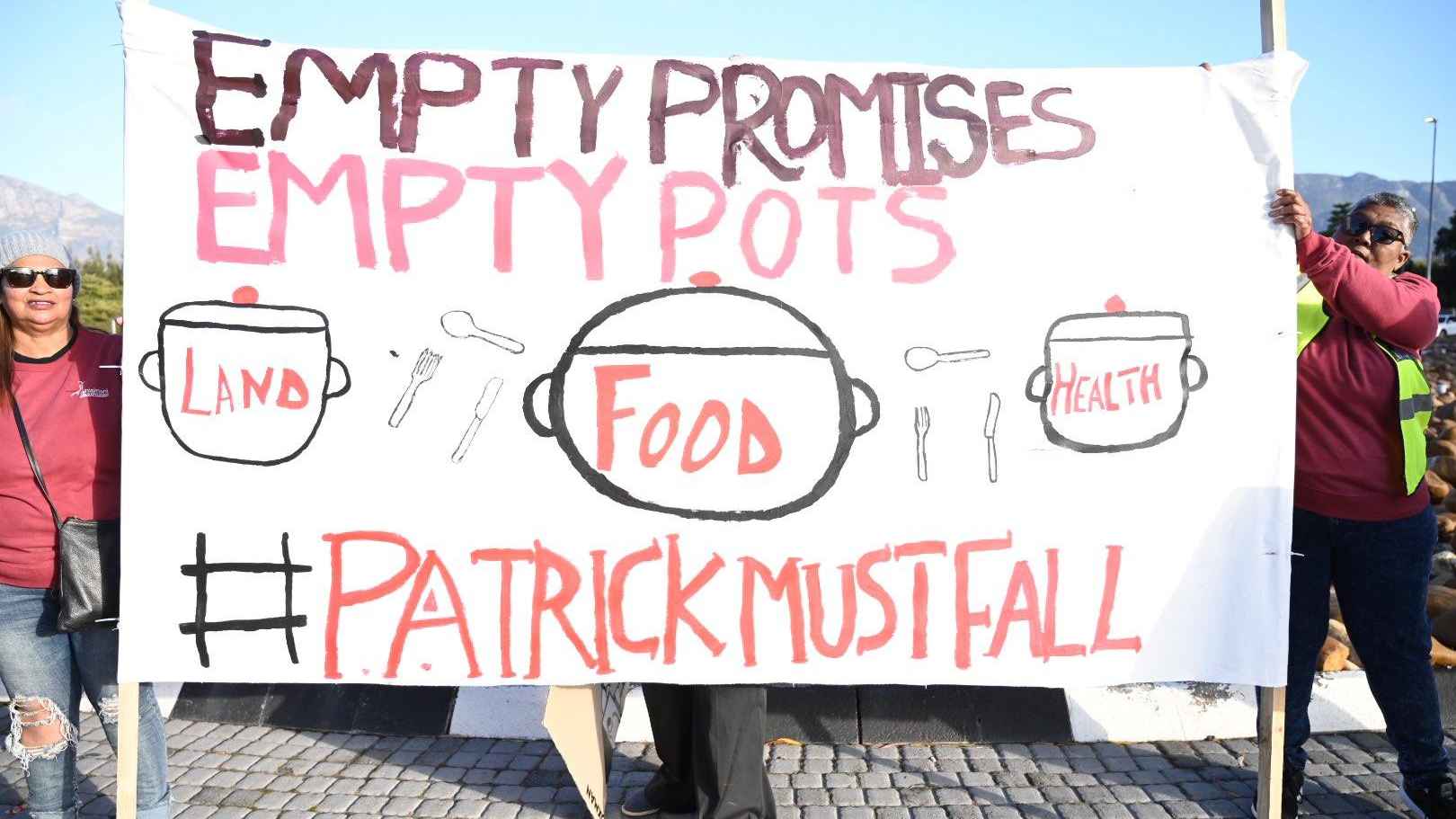 An image of a protest by women holding a board that says Empty Promises, Empty Pots, hashtag Patrick Must Fall